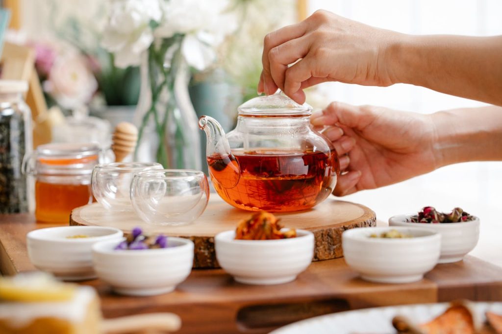 A person's hands preparing a glass teapot of amber herbal tea surrounded by small bowls of dried Ayurvedic herbs, honey, and glass cups on a wooden tray.