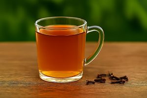 A warm, dark amber-colored herbal tea in a clear glass mug with whole dried cloves scattered on the wooden table beside it.